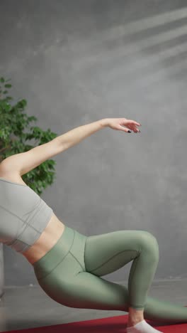 vertical video confident and athletic brunette girl in a gray top and green sweatpants does yoga while sitting on a red sports carpet and stretches her hand up near infected candles and a house plant at home