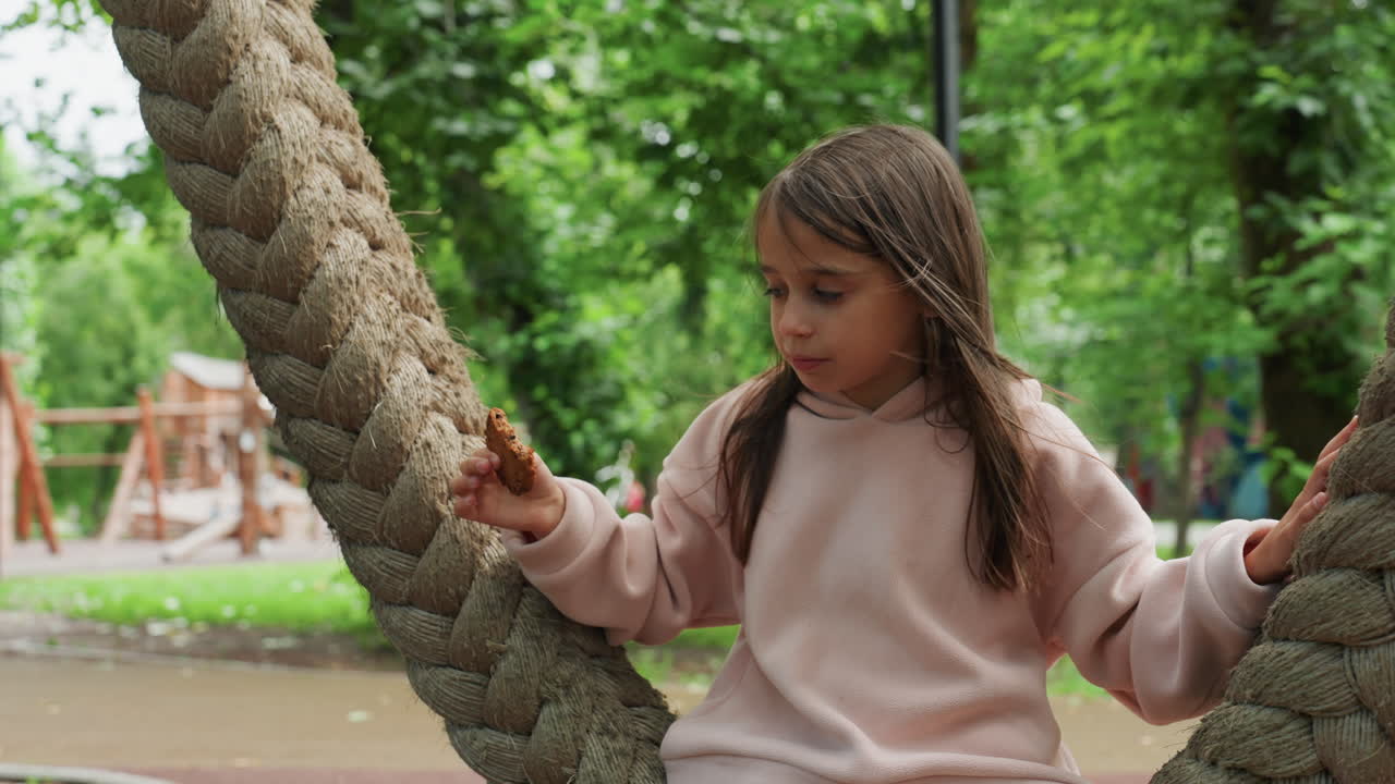Niña saboreando un dulce. Niña disfrutando una galleta al aire libre. Niña con cabello trenzado prueba un dulce cerca del columpio del parque. Niña con suéter rosa y cabello trenzado disfruta tranquilamente una galleta recién horneada al aire libre.