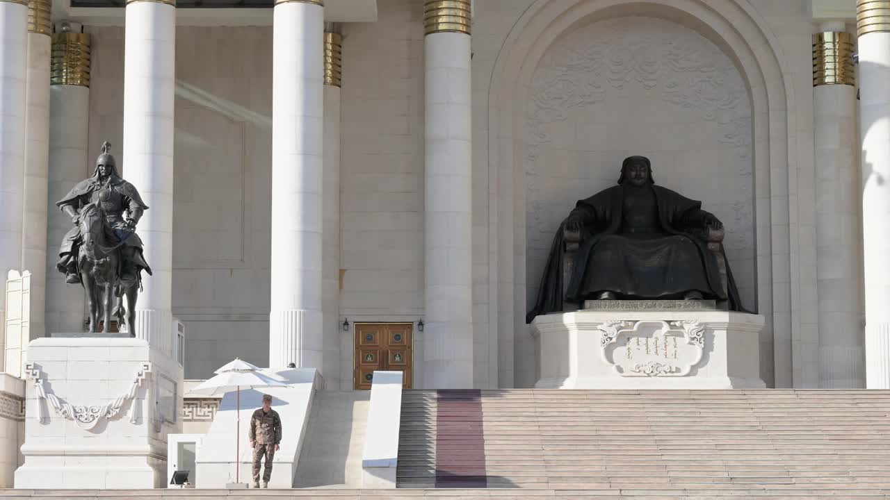 A wide-angle view of a Mongolian soldier standing guard beside the bronze statue of Genghis Khan at Sukhbaatar Square, with the Government Palace in the background, on a sunny day.