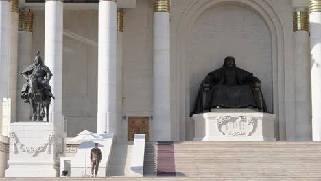 a wide-angle view of a mongolian soldier standing guard beside the bronze statue of genghis khan at sukhbaatar square, with the government palace in the background, on a sunny day.