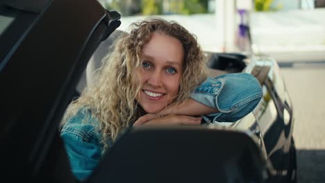 Beautiful-blonde-girl-with-blue-eyes-posing-and-looking-at-the-camera-while-sitting-in-the-front-seat-of-a-dark-gray-cabriolet