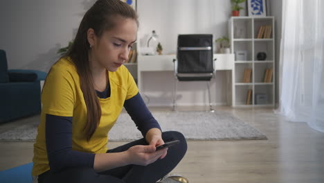 Una-Joven-Morena-Con-Un-Teléfono-Inteligente-Descansa-En-Una-Habitación-Deportiva-En-Un-Apartamento-Sentado-En-Una-Alfombra-Usando-Redes-Sociales-E-Internet,-Un-Estilo-De-Vida-Deportivo-Y-Saludable