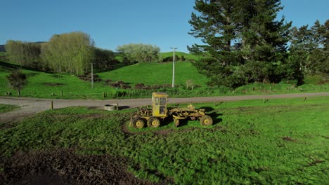 viejo grader amarillo ocioso en el campo de la granja rodeado de colinas y árboles bajo un cielo azul claro.