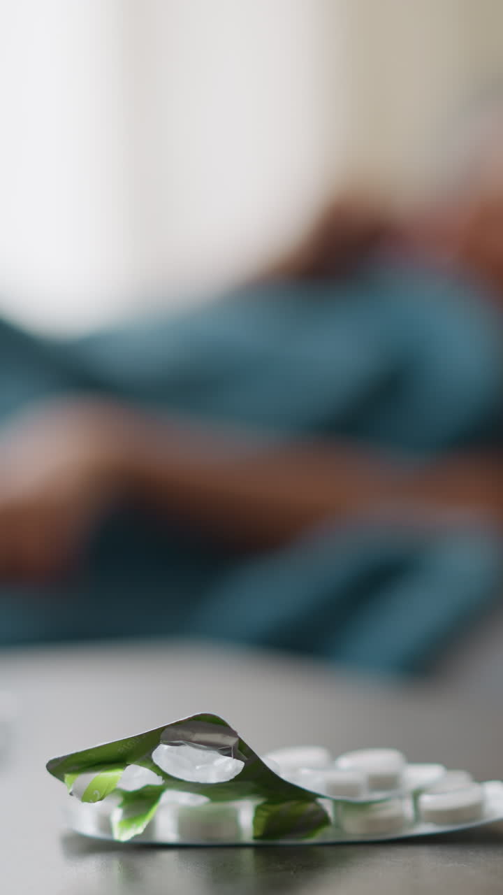 Nærbillede af medicin og glas på bord med sløret udsigt til person, der justerer sig i sengen, hvilket antyder sygedag, bedring og komfort i et fredeligt indendørsmiljø