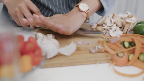 midsection of biracial woman in apron preparing meal, crushing garlic clove in kitchen, slow motion