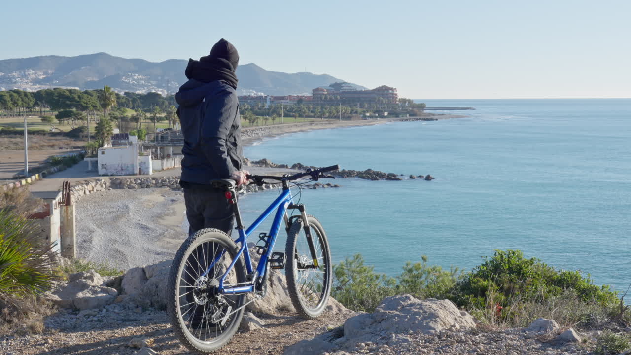 Traveller walks on rocky trail with mountain bike, stops to admire ocean view, then lifts bicycle on his sholder and walks towards coastal city