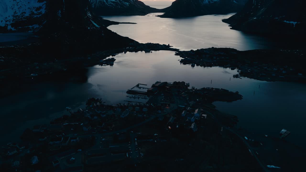 Letecký pohled za soumraku na Reine, Norsko, se zasněženými horami obklopujícími fjord a vesnici podél pobřeží. Záběr s náklonem nahoru.
