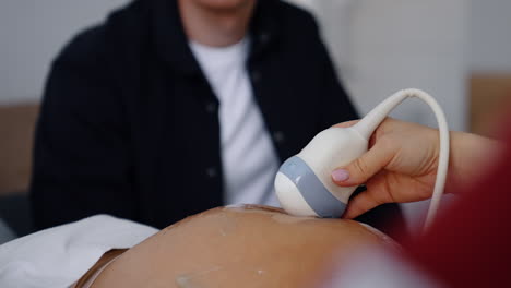 female hand of a doctor holds the ultrasonic device on round belly of pregnant. cropped image of man sitting at backdrop.