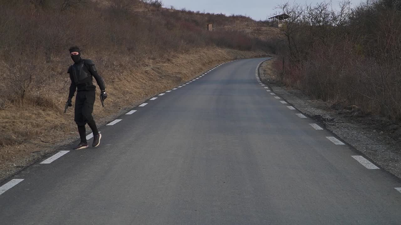 A mysterious ninja in black armor walks cautiously on an empty rural road, gripping dual weapons. The scene exudes suspense and action, perfect for martial arts, stealth, and cinematic projects