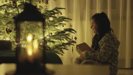 a girl seated next to the christmas tree, holding a gift and gently touching the hanging decorations - close up