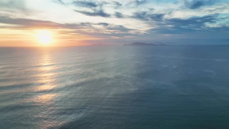 Drone-shot-panning-up-from-open-blue-ocean-and-gentle-waves,-to-golden-sunset-and-distant-island-on-the-horizon