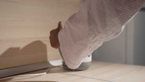 close-up of man in bathrobe gripping electric kettle with one hand, preparing to pour water. soft morning light casts shadows on minimalist kitchen surface, highlighting sleek kettle design