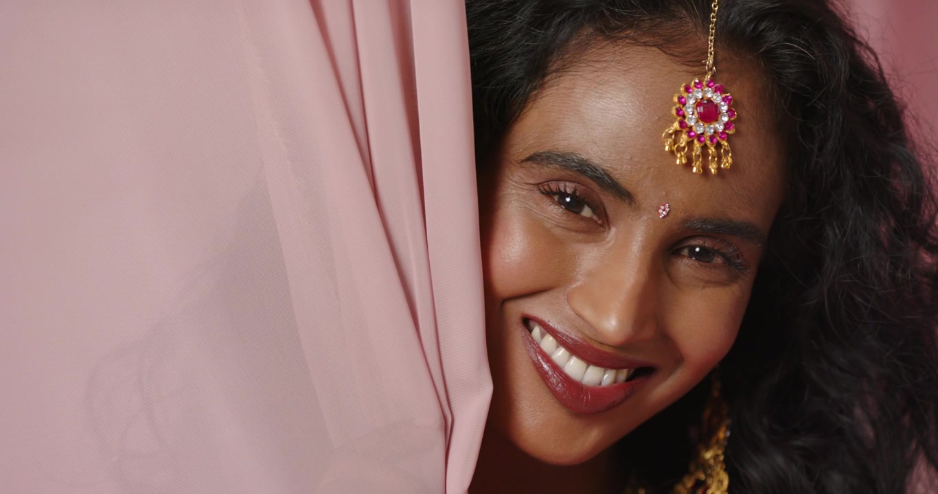 Adult Indian woman on camera cue smiling, lifting pink drape at studio peeking showing gold jewelry