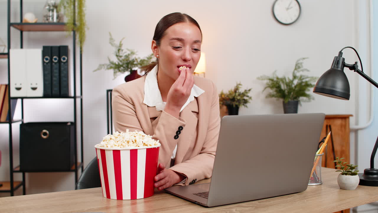 Young businesswoman enjoys popcorn during break launches series trailer on laptop and giggles today