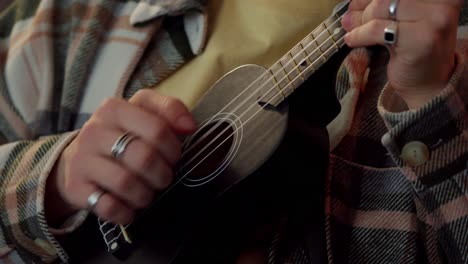 cerrar un tipo con una camisa a cuadros con sus manos con anillos juega un ukulele negro en casa en una atmósfera acogedora