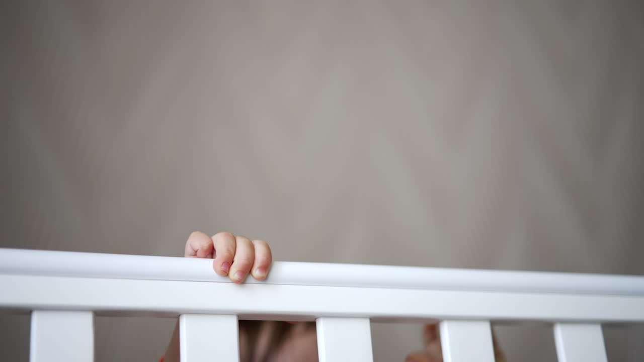 Cheerful happy baby boy playing hide-and-seek in his crib. Lovely laughing baby sits and stands up in white cot. Close up.
