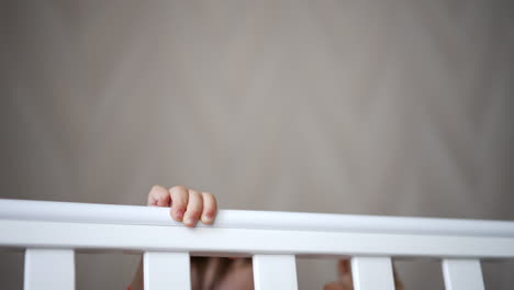 cheerful happy baby boy playing hide-and-seek in his crib. lovely laughing baby sits and stands up in white cot. close up.
