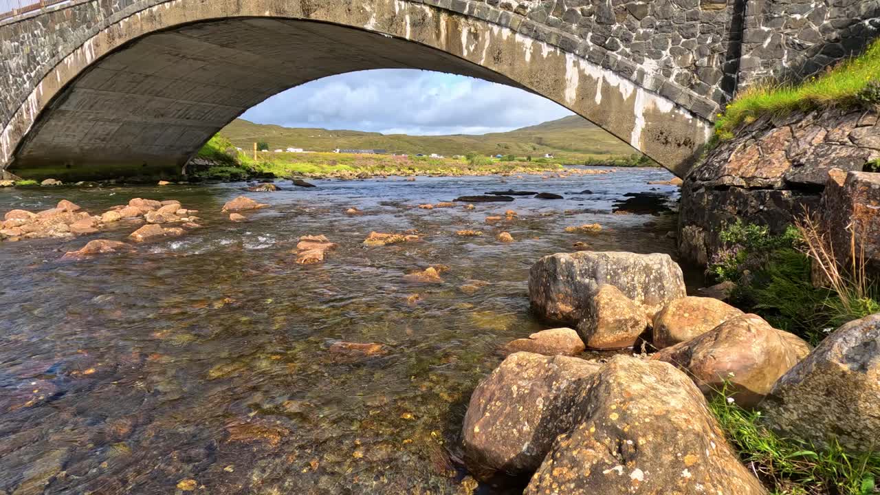 Stone arch bridge crosses clear rocky stream with lush green banks in rural daylight landscape