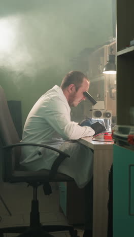 una vista de cerca de un técnico en una bata de laboratorio blanca trabajando diligentemente en componentes electrónicos bajo un microscopio con la luz encendida detrás de él y un estante junto a la entrada
