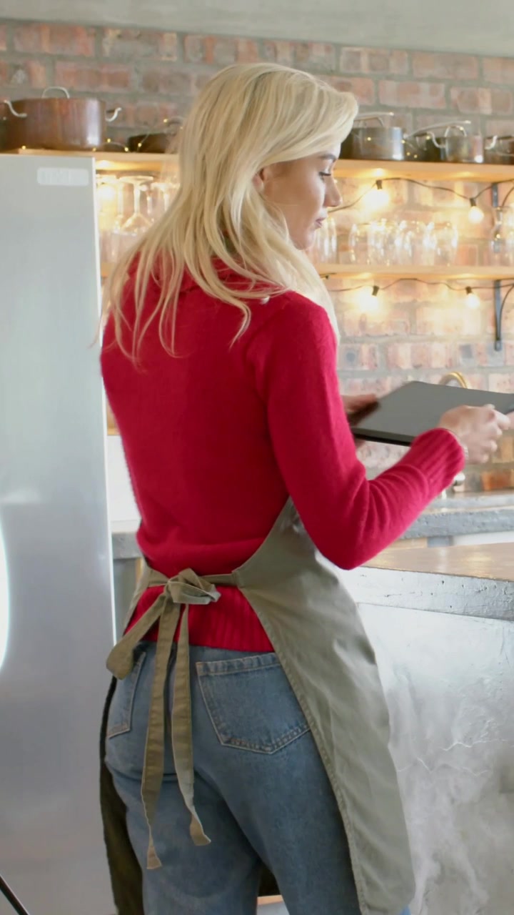 Vertical video: Picking up tablet, woman in olive apron adjusting ring light to record in kitchen