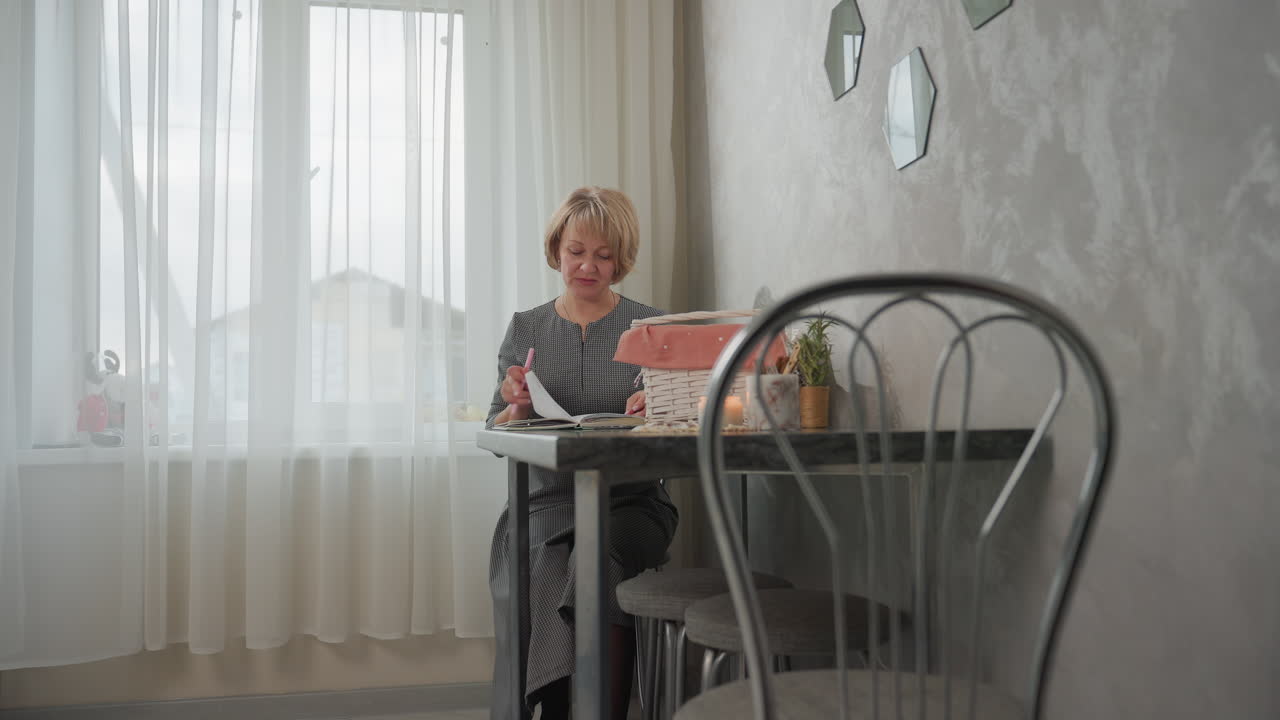Student seated at table adjusting while opening book in hand holding pen near basket candle and plants on cozy table in softly lit room with decorative wall mirrors and sheer curtains by window