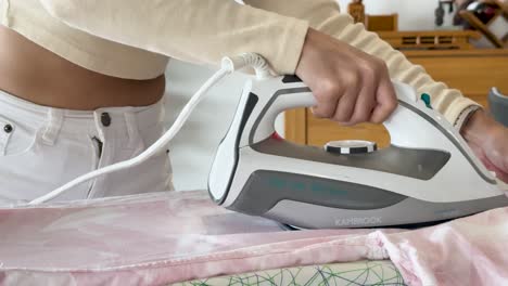 a young woman uses a steam iron to press a pink shirt on an ironing board in a bright, modern home interior with natural daylight