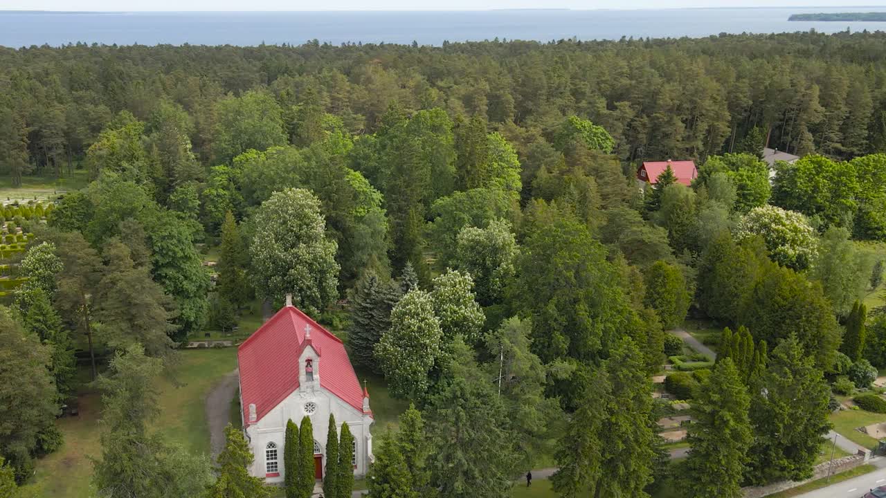 Aerial drone footage orbiting and spinning around a rural countryside gorgeous white church with red roof that is in a sunny forest while Baltic sea ocean is visible in the horizon at background.