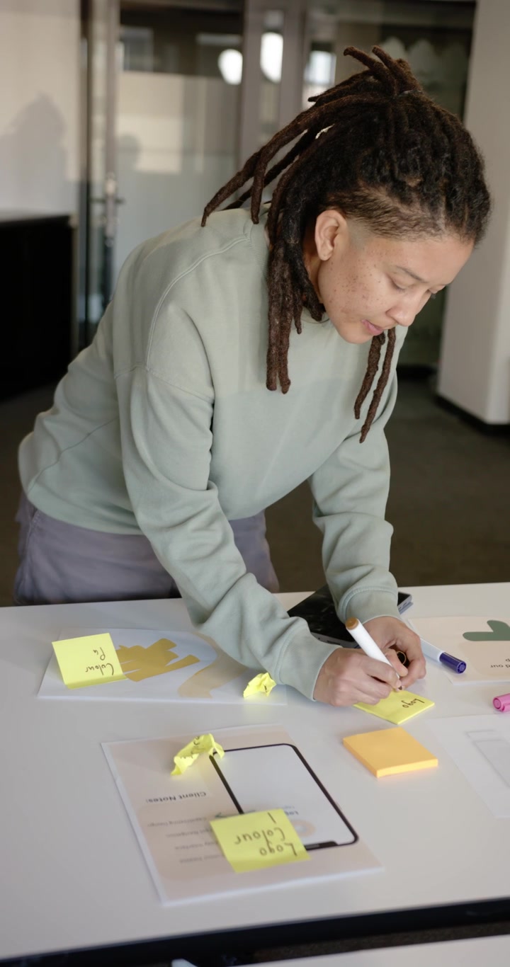 Video verticale: Persona afroamericana non binaria che si china per prendere appunti su mockup per il design in ufficio.