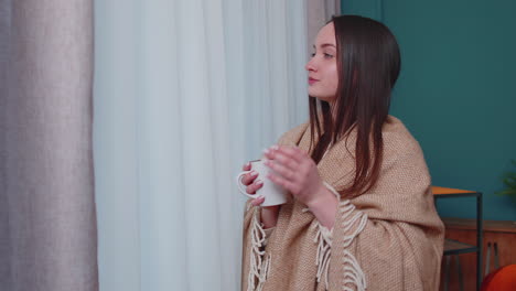 Young-adult-brunette-woman-in-warm-white-sweater-drink-hot-beverage-mug-relaxing-at-home-near-window