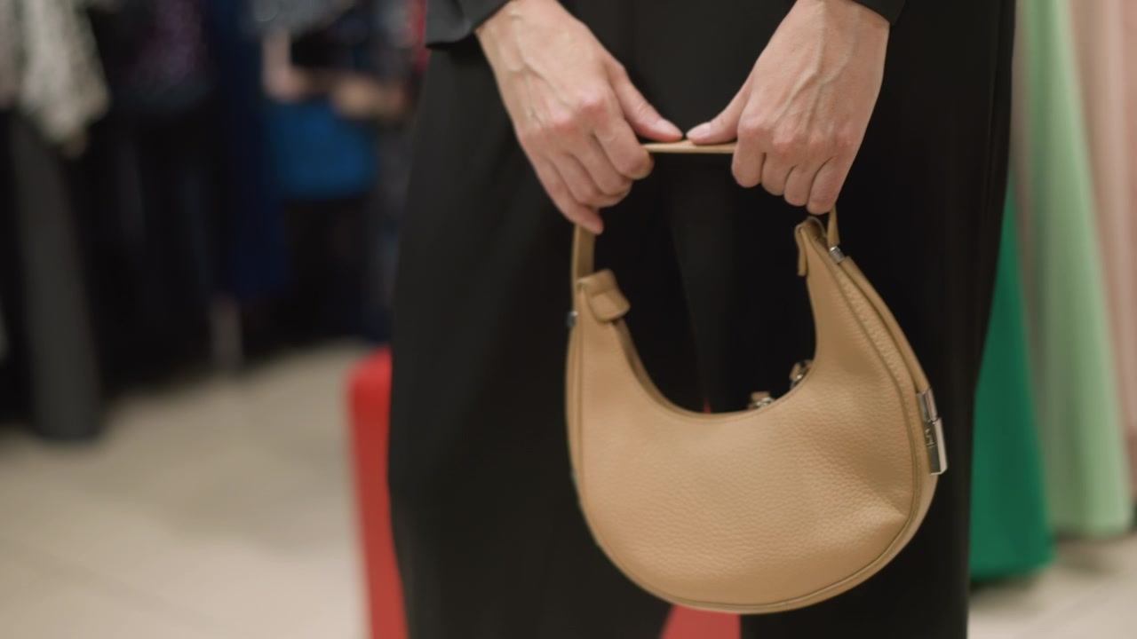 Primo piano di una donna che tiene ed esamina una borsa beige in luce soffusa. La texture della borsa, la forma e gli accessori in argento sono visibili mentre il soggetto ne regola la posizione contro l'abbigliamento. Appendiabiti.