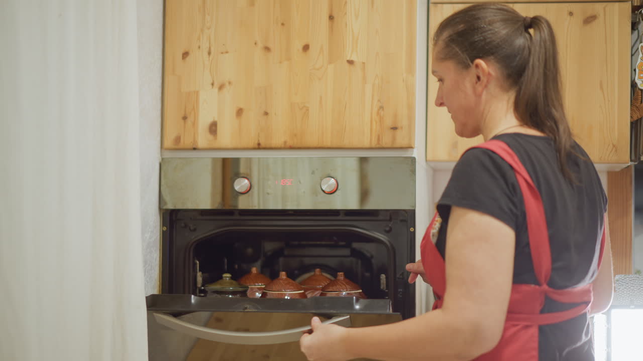 Mujer blanca cocinera casera retirando bandeja de asado del horno, guantes de cocina en las manos, anfitriona sonriente presentando asado de cerámica en bandeja, vapor y guarnición, acogedora escena de cena nocturna, hospitalidad y satisfacción.