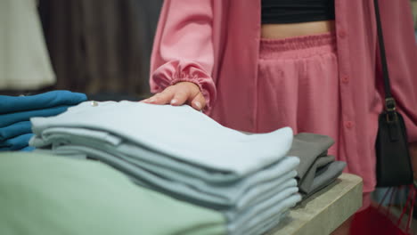 vista inferior de una mujer con una camiseta negra bajo un traje rosa, llevando bolsas de compras, tocando y sintiendo suavemente la textura de la ropa bien plegada en la mesa mientras las observa en una tienda de ropa