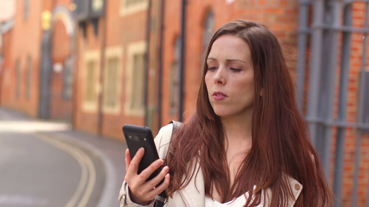 Woman Outdoors Walking To Work Along City Street Lined With Houses Looking At Mobile Phone 1
