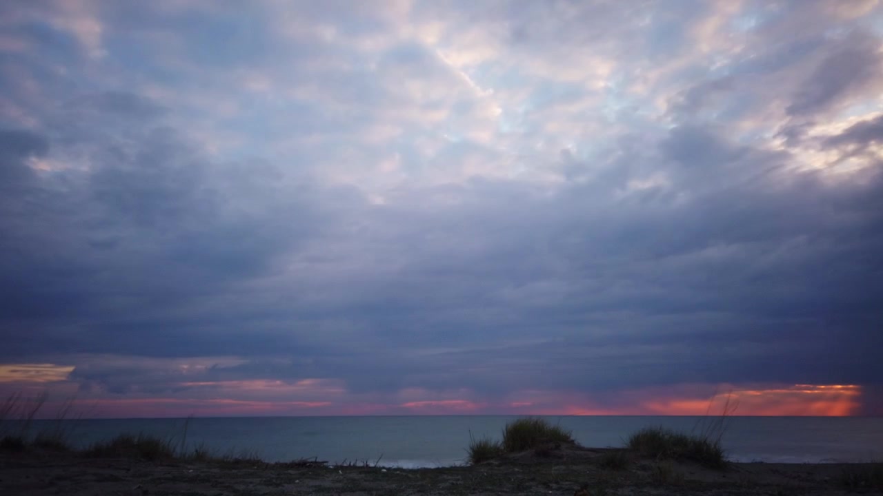 Wide landscape time-lapse from pitch darkness to dawn over sea with fast-moving dark overcast clouds revealing soft pink sunrise light on horizon.