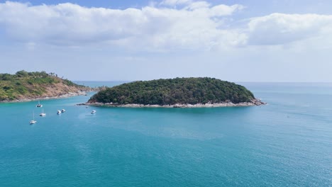 Drone footage captures serene island bay with boats in Phuket, Thailand. Clear skies and turquoise waters create a tranquil scene