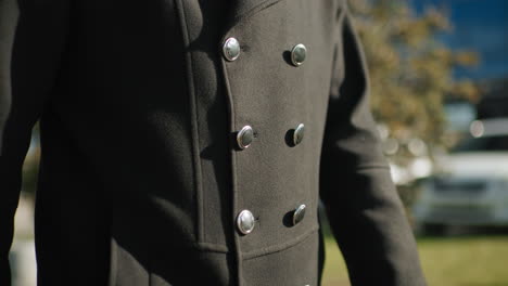 close-up of elegant black coat featuring shiny metallic buttons in an outdoor urban setting with blurred background, including parked cars and greenery