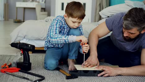 curious child is driving screw in piece of wood with screwdriver and his dad is holding wooden sheet and teaching him basics of woodwork. united family concept.