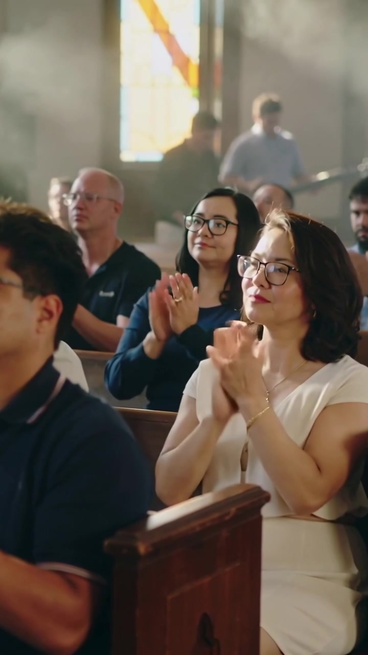 Video verticale: Uomo con polo blu scuro che applaude in chiesa rispondendo al fronte, raggi di sole, altri che si uniscono.