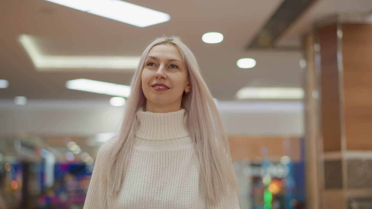 back view of mall explorer walking under bright decorative lights smiling and admiring crystal decor while wandering through modern retail corridor wearing white knit sweater and pastel hair flowing