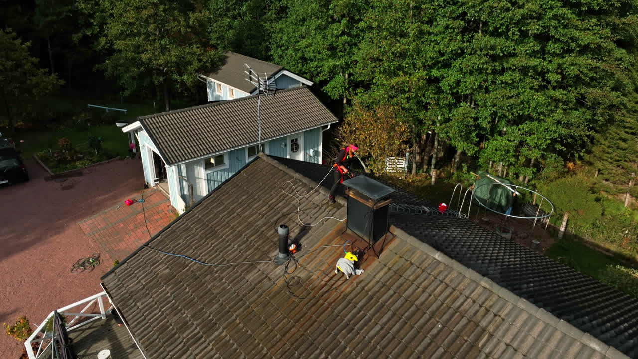 Drone descending in front of a woman pressure washing a house roof, fall day