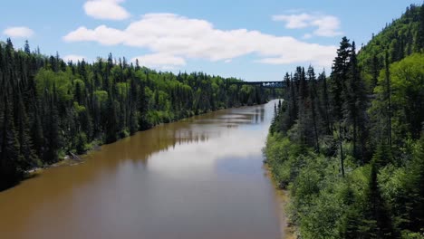 a drone flies down down a river surrounded by forest on both sides and old railroad bridge can be seen in the background on a bright cloudy sunny summer day