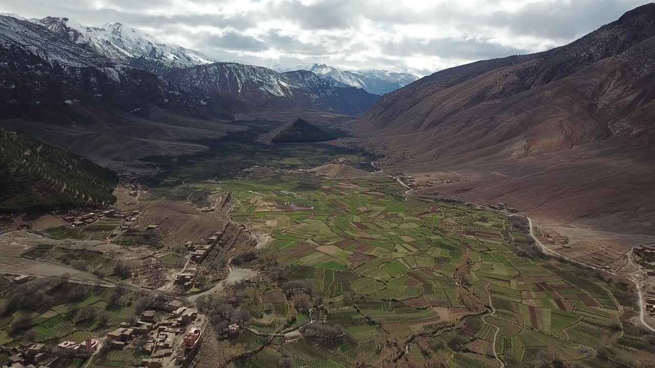 A magnificent valley cuts through Morocco's High Atlas, where towering peaks frame lush green fields, ancient kasbahs dot the landscape, and mountain rivers nourish the fertile earth