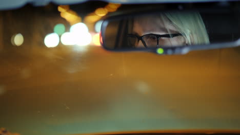 Una-Mujer-Con-Gafas-Conduce-Un-Automóvil-Por-La-Ciudad-De-Noche-Su-Rostro-Se-Refleja-En-El-Espejo-Retrovisor-4