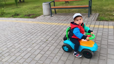 adorable toddler riding happily by the park. baby boy in cap drives toy car. top view.