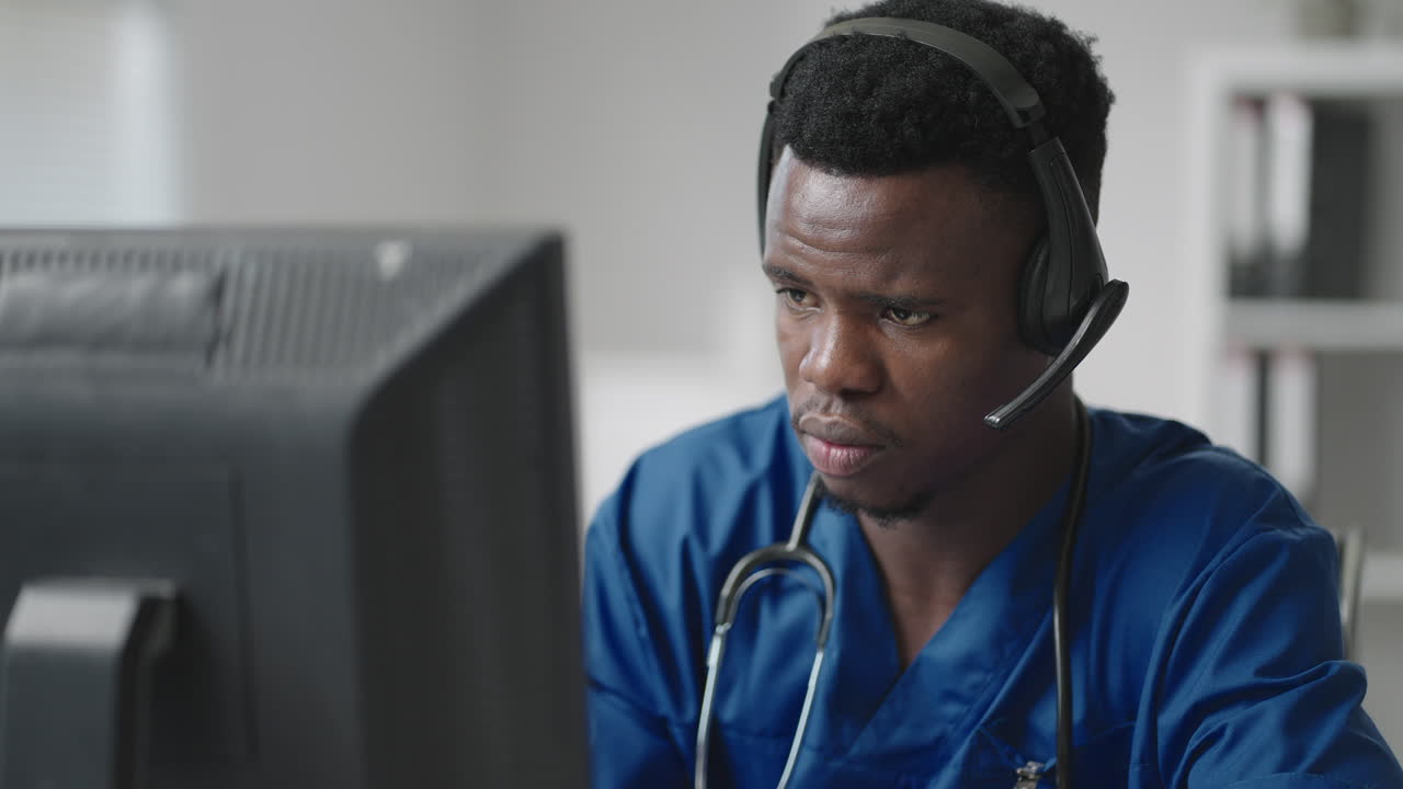 A black man sits at a computer in a doctor's uniform and writes a patient's card while taking calls with headphones. Ambulance Hotline receive calls and distribute ambulances