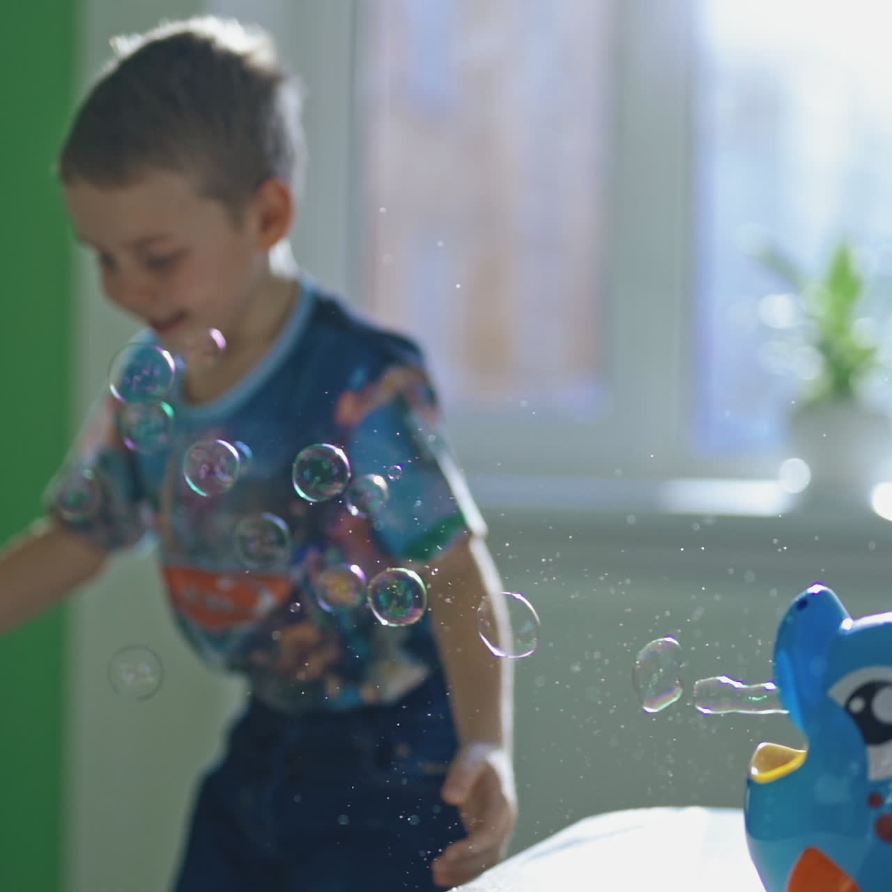 Little boy is popping soap bubbles indoors. Curious child catches circle bubbles at home. Bubble blower in the form of a fish and a small kid plays happily nearby.