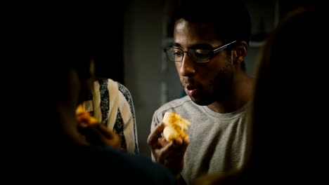 joven afroamericano comiendo pizza en una fiesta casera. los jóvenes disfrutan de comida rápida en la cocina de casa