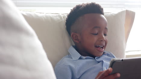 Happy-african-american-boy-lying-on-sofa-using-tablet-at-home,-slow-motion