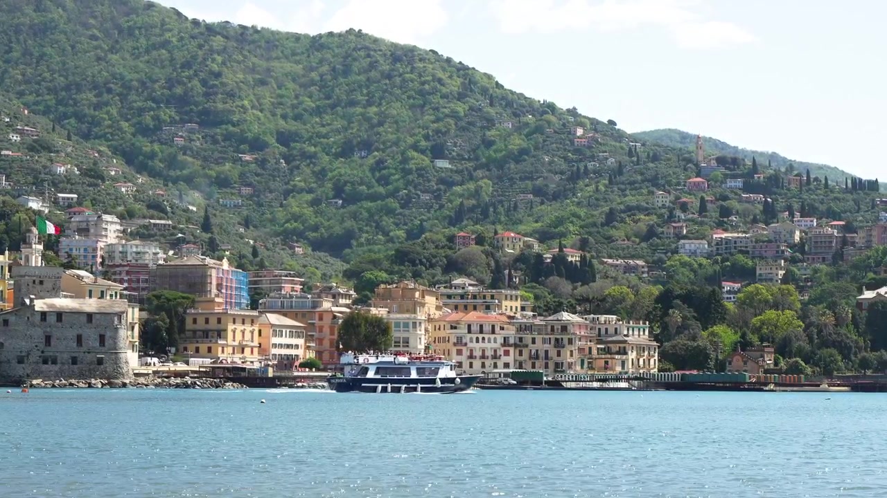 Uno scatto panoramico suggestivo cattura il Castello di Rapallo e i vivaci edifici sul lungomare lungo la Riviera Ligure, con barche turistiche sull'acqua azzurra, rigogliose colline verdi e la luminosa luce del giorno, perfetto per il viaggio o per