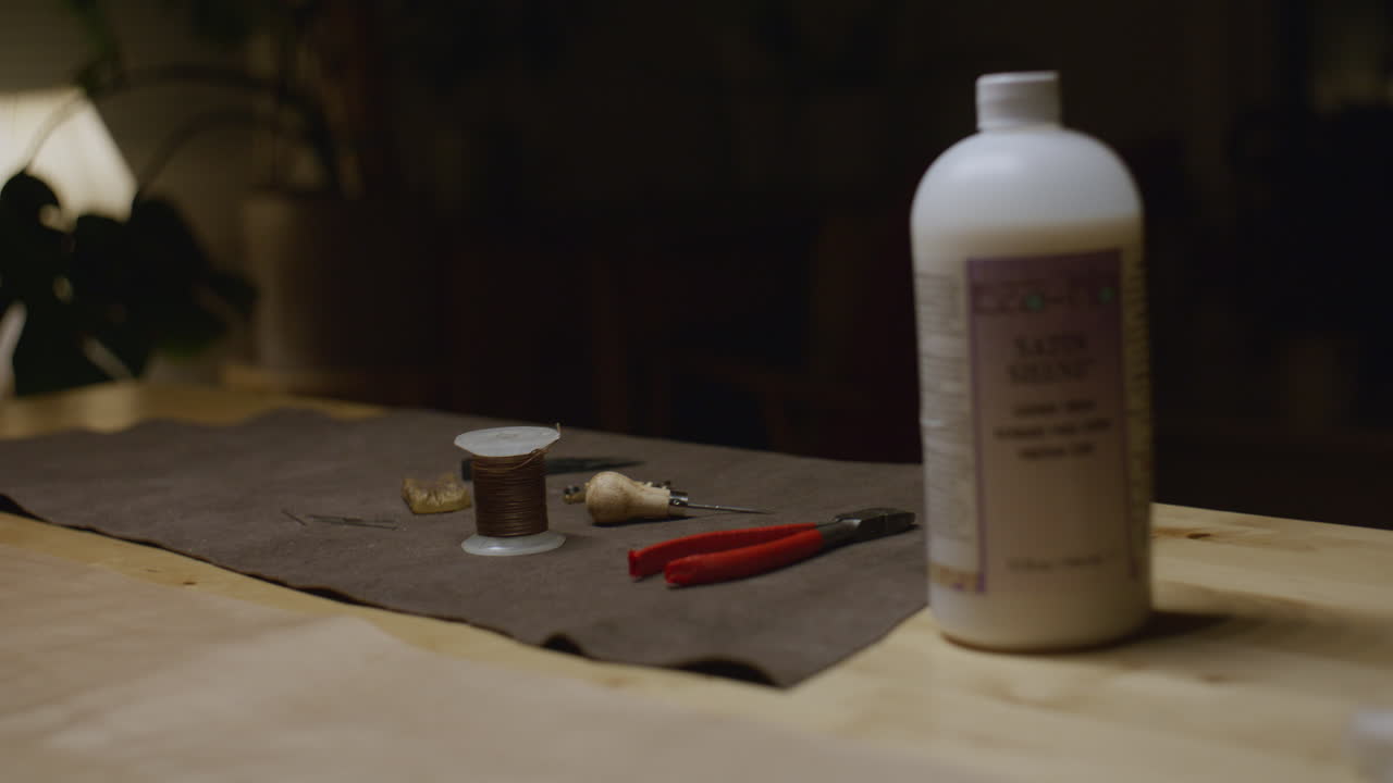 A set of leatherworking tools, including a spool of thread, awl, pliers, and a bottle of finishing solution, arranged on a brown suede mat atop a wooden workbench.